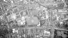 A historic black and white vertical aerial photo of the city of Dublin. To the left are several groups of buildings arranged around courtyards. They form the shape of a Greek cross. To their left are several playing fields. 