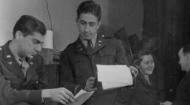 A historic black and white photo of two men with dark hair in US Air Force uniforms consulting some paperwork. One man holds a smoking cigarette. In the background a third man and a woman are talking.