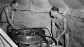 A historic black and white photo of 2 men in shorts and with their shirts off processing photographs using metal tanks in a tent. 