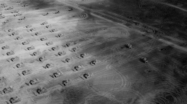 A historic black and white oblique aerial photo of a dusty landscape. The landscape is marked with numerous vehicle tracks. Arranged across it in a grid formation are tanks, each accompanied by a pair of soldiers.