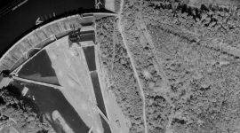 A historic black and white vertical aerial photo of a dam. The dam is to the left of the image, holding back a large body of water. To the right is a large wooded area.