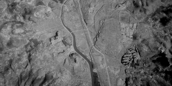 A black and white vertical aerial photo of a city. A river runs through the middle of the image with an oval racetrack to its right. A few buildings are visible, but most have been destroyed. Hills are visible either side of the built-up area.  