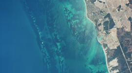 A historic colour vertical aerial photo of a coastline. The left two-thirds of the image are filled with a very blue sea. The shore to the right looks dry and arid. 