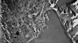 A historic black and white vertical aerial photo of a bay surrounded by mountains. A small cluster of buildings is visible with ships moored offshore. Snow covers some of the mountains. 