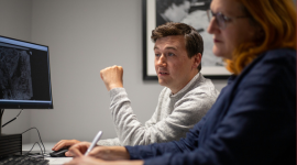 A man sits looking at a computer screen, using a computer mouse. In the foreground, out of focus, a woman with long red hair makes notes.