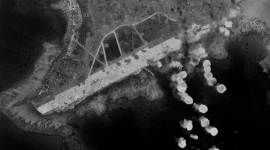A historic black and white vertical aerial photo of a runway on an island. The runway stands out white against the forested land and darkly coloured sea. To the right of the image, numerous white clouds indicate where bombs are exploding.