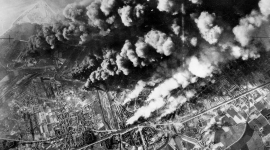 A historic black and white vertical aerial photo of a town. Thick columns of black, grey and white smoke rise from fires across the town. The sea is just visible in the top left corner. 