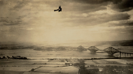 A historic black and white photo of 3 men standing on a rocky outcrop above the sea, using a surveying instrument on a tripod. 