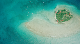 A historic black and white vertical aerial photo of a bay surrounded by mountains. A small cluster of buildings is visible with ships moored offshore. Snow covers some of the mountains. 