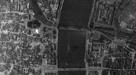 A black and white vertical aerial photo of the city of Cologne. The cathedral is clearly visible on the left bank of a wide river. The river is spanned by a railway bridge and a road bridge. Buildings either side of the river show clear signs of bomb damage. 
