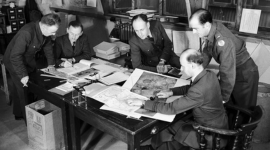 A black and white historic photo of 5 men in uniform around a table. They are studying various photos and maps. 2 pocket stereoscopes are also visible. 2 of the men are seated, 2 stand and 1 leans on the table. 3 of them are smoking. 