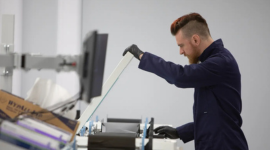 A man with a beard and red mohawk operates a flatbed scanner. He is wearing a blue lab coat and black gloves. One hand lifts the lid of the scanner while the other rotates the film spool set over the scanner. A computer monitor and some boxes are out of f